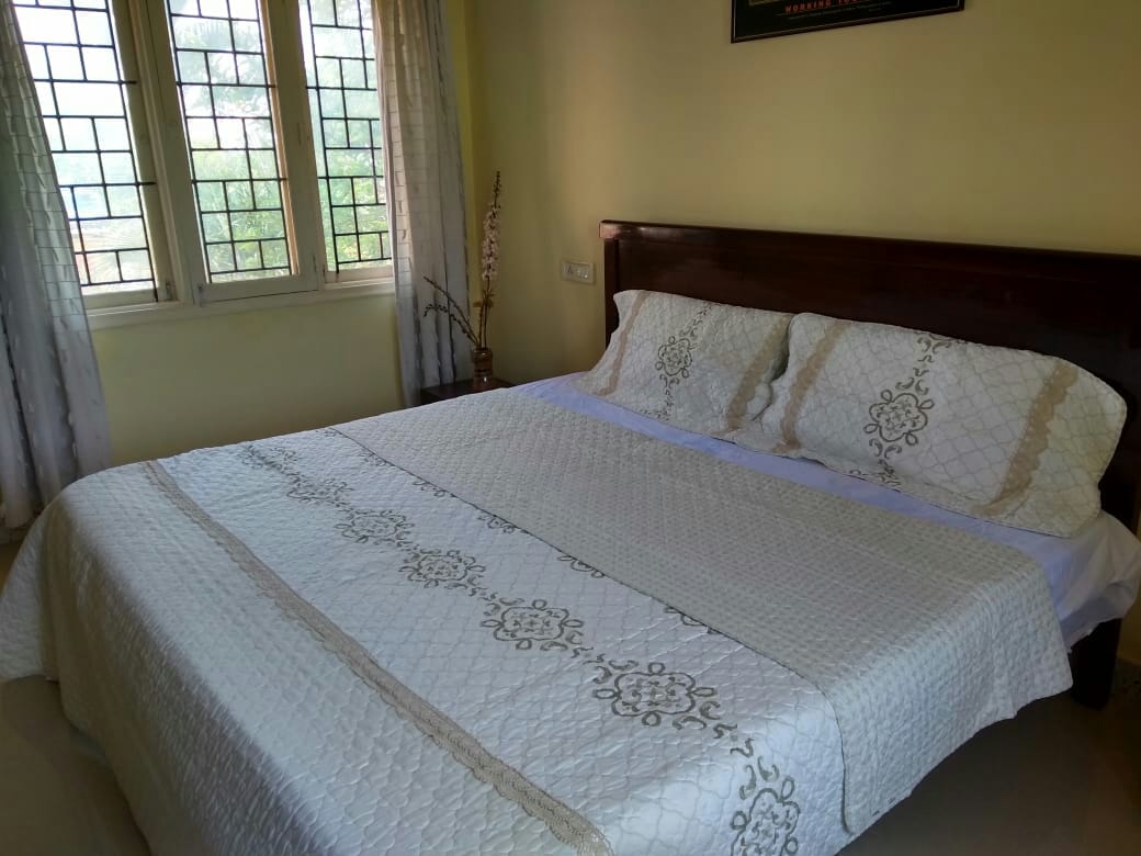 A spacious bedroom is presented, featuring a large bed draped with a light-colored quilt. Two windows with decorative panes allow natural light to fill the room, while soft curtains frame the view of the outside greenery.