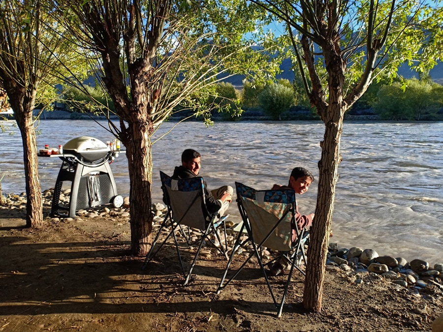 Two comfortable folding chairs are positioned near a flowing river, under the shade of trees. A barbecue grill stands nearby, set against a backdrop of natural greenery. The gentle movement of the water and the tranquil environment create a peaceful outdoor setting.