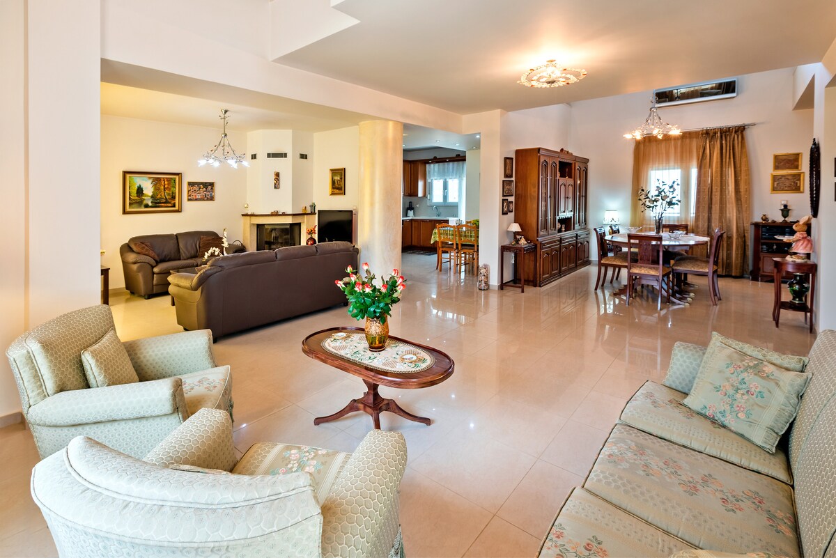 A spacious living area is presented with elegant furniture, including a pair of soft armchairs and a wooden coffee table. A dining table is visible in the background beside a cozy seating arrangement. Soft lighting from chandeliers enhances the inviting ambiance while natural light streams through large windows.