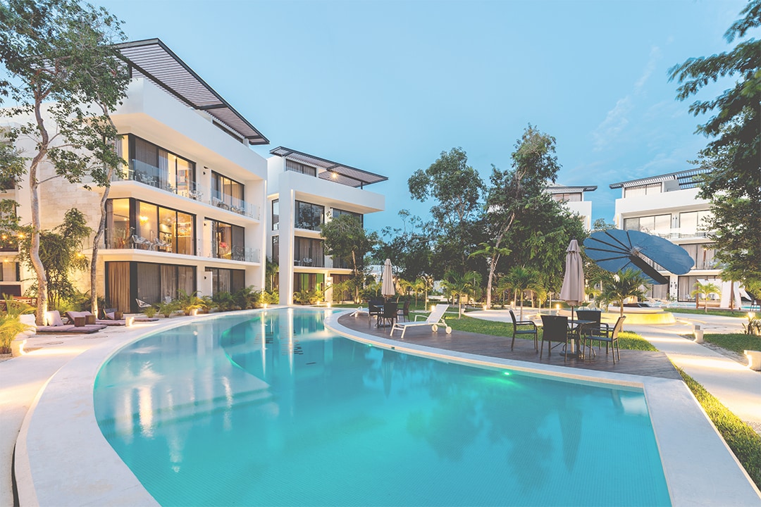 A serene pool area is surrounded by modern buildings, showcasing gentle curves and lush landscaping. Comfortable lounge chairs and umbrellas are positioned along the pool's edge, while dining tables and chairs are set nearby for outdoor relaxation.