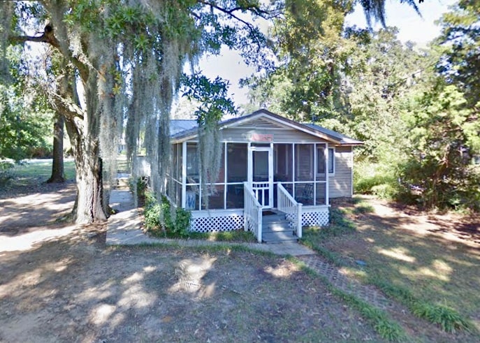 A charming cottage is surrounded by a grassy area and mature trees, including a large oak draped in Spanish moss. The screened-in porch is accessible via a set of steps, offering a view of the natural surroundings.