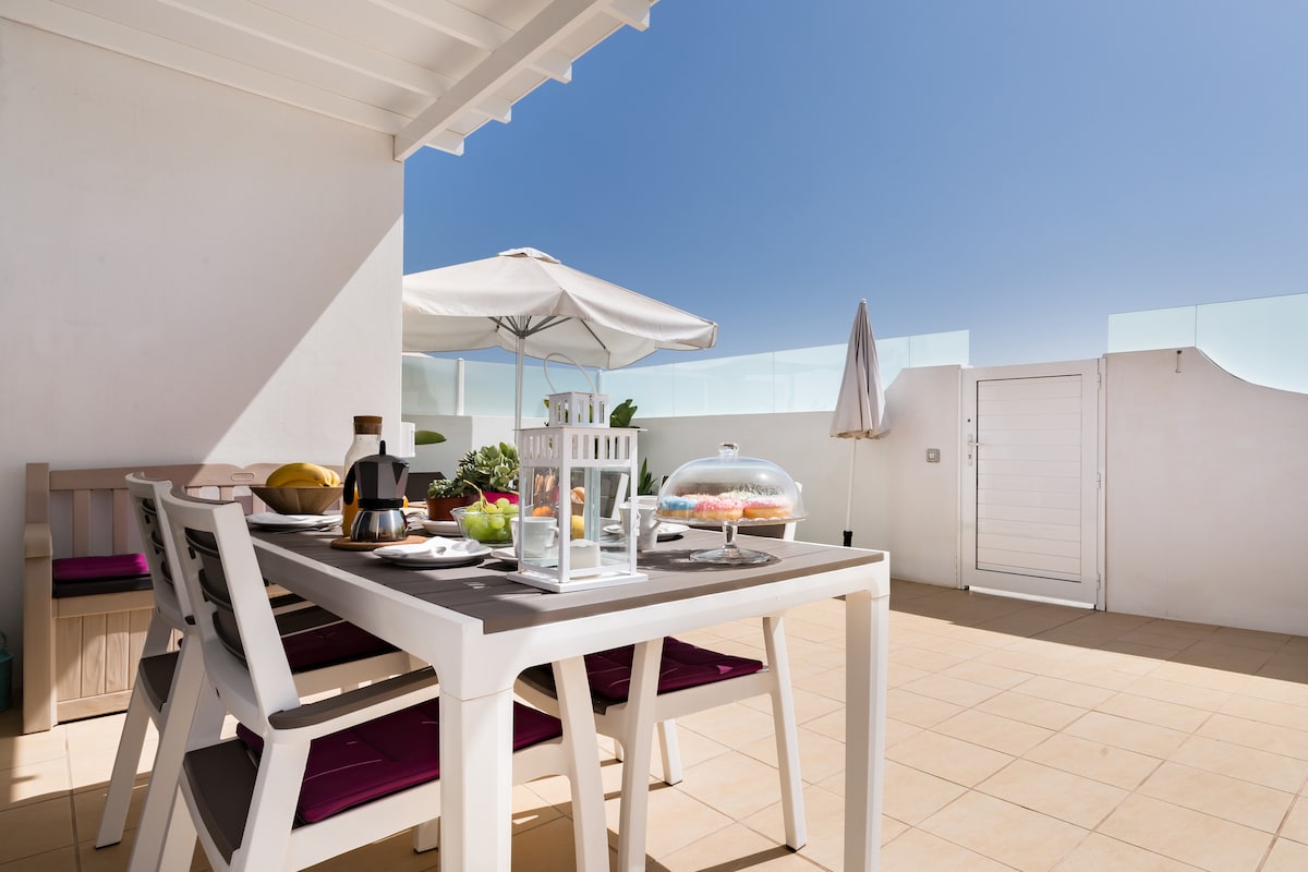 A spacious outdoor terrace is presented, featuring a white dining table set with plates of fresh fruit and pastries. An umbrella provides shade, while a glass dome covers a dessert. The bright blue sky completes the inviting outdoor space.