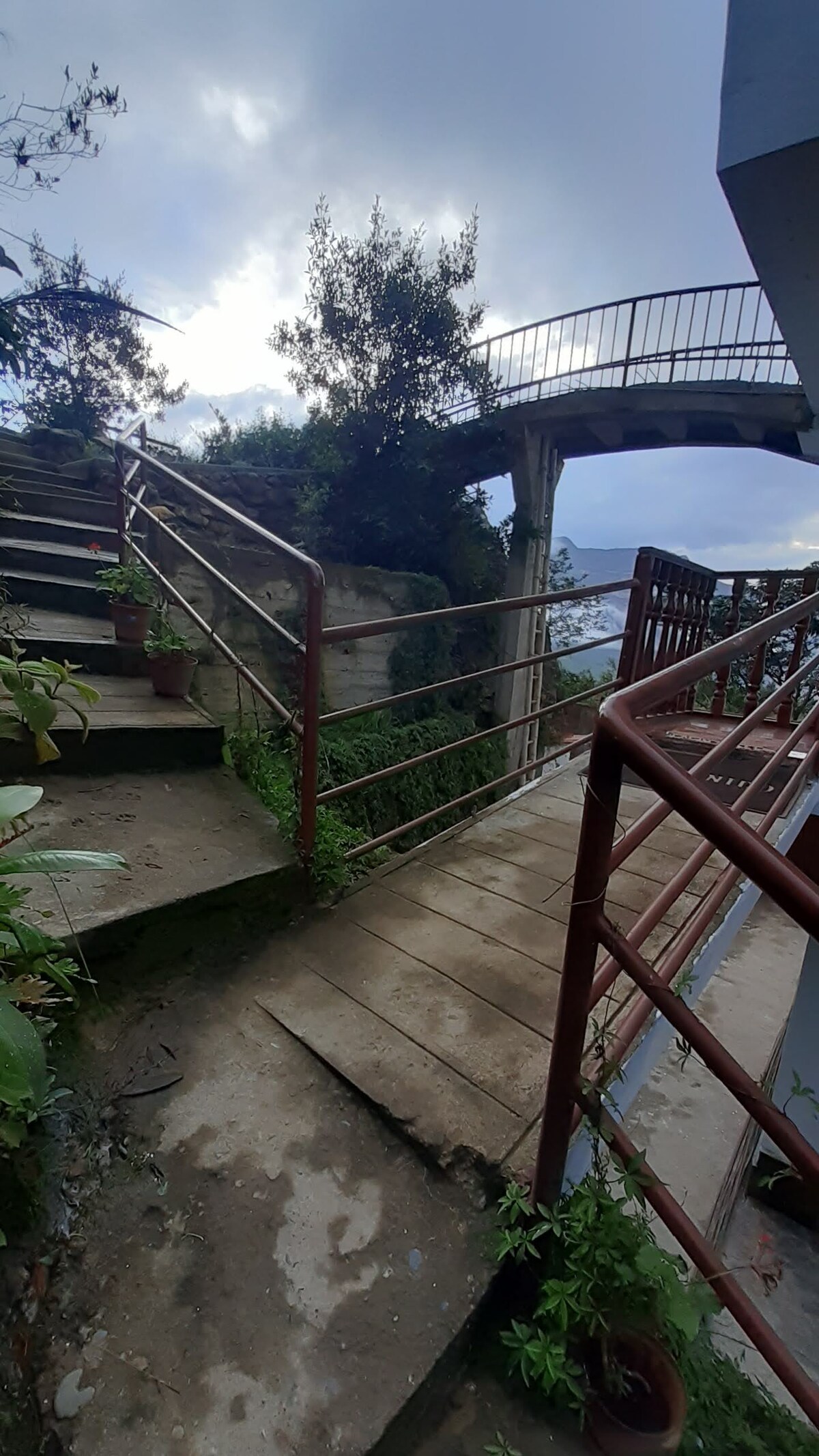 A stairway with metal railings leads upwards, surrounded by lush greenery. An adjacent pathway connects to a bridge above, offering views of the natural landscape. The sky features soft, diffused light indicating early morning or late afternoon.