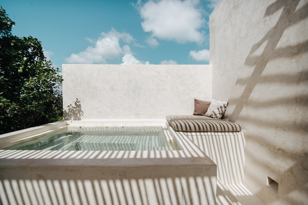 A private rooftop pool is depicted, surrounded by smooth, white walls that reflect natural light. A comfortable cushioned lounge area is positioned beside the pool, with shadows cast by nearby structures. The clear blue sky and scattered clouds create a serene backdrop.