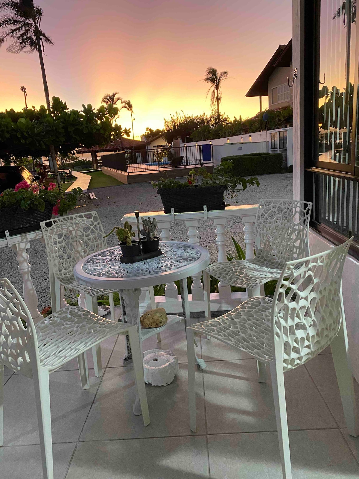 An outdoor seating area is set with a round table surrounded by four intricately designed white chairs. A small potted plant rests in the center, and the background features a colorful sunset casting warm tones across the sky.