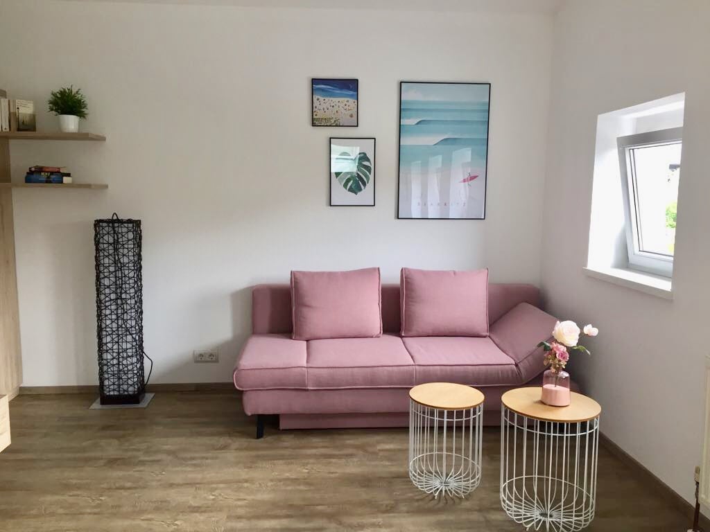 A modern living space features a pink sofa with a sectional design, complemented by two round side tables. Decorative wall art enhances the aesthetic, while a small plant adds a touch of greenery. Natural light filters through the window, highlighting the neutral color palette.
