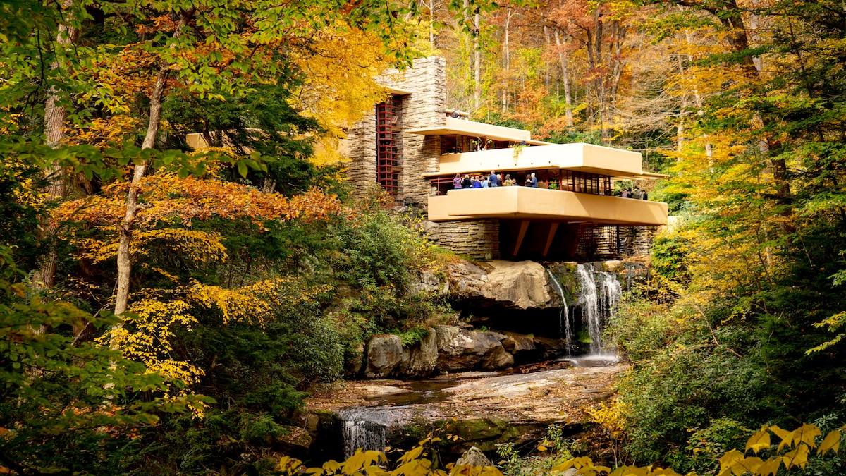 A modern structure is nestled among vibrant autumn foliage, with a waterfall cascading nearby. The design features layered levels, allowing for ample outdoor seating that blends with the surrounding nature. The warm tones of the leaves contrast with the building's neutral colors, creating a harmonious scene.