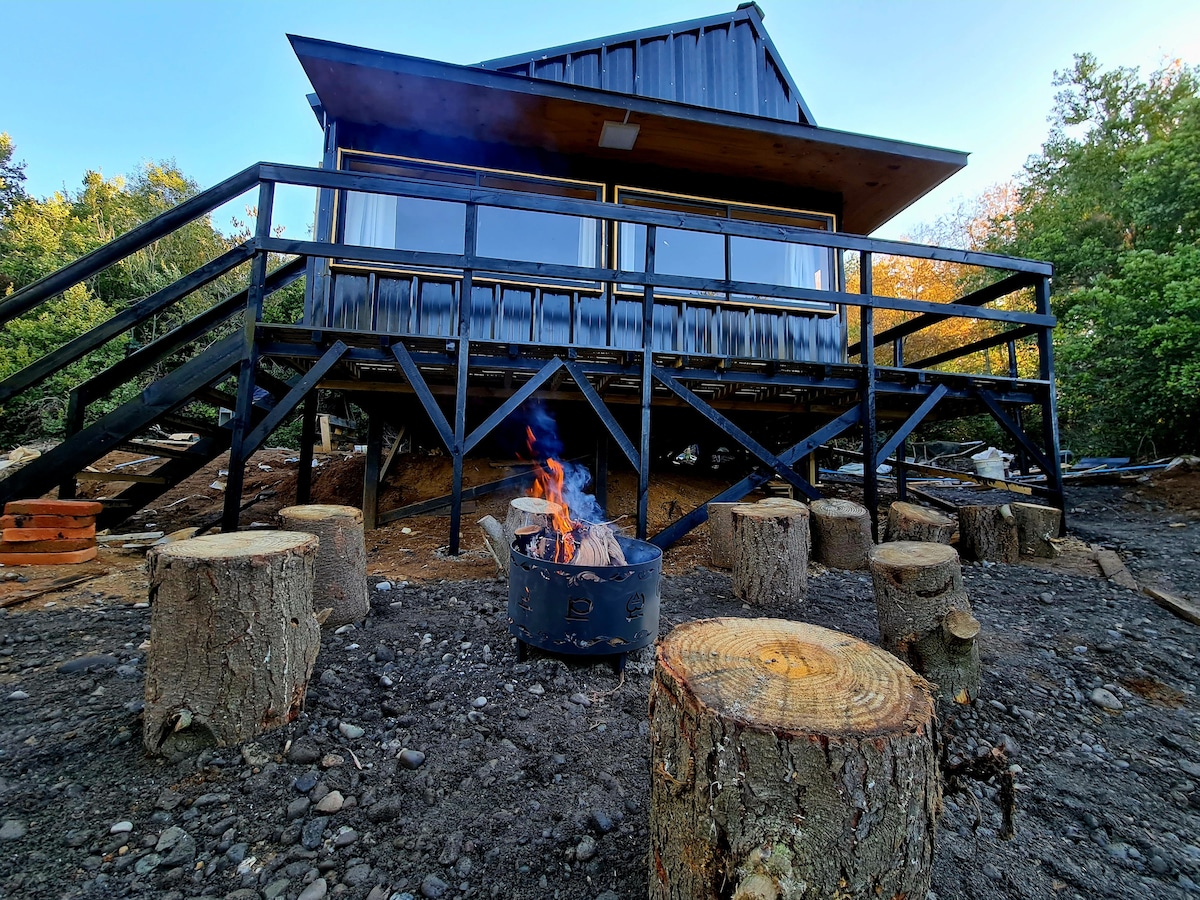 An outdoor seating area is shown with log stools arranged around a fire pit, where flames flicker above glowing embers. The cabin's exterior features large glass windows and a wooden deck, surrounded by greenery, creating a connection to the natural environment.