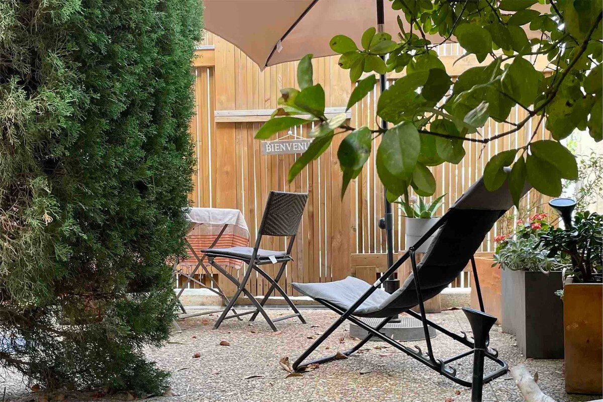 A private patio space is surrounded by greenery, featuring a lounge chair and a small table with chairs. The area is shaded by a large umbrella, offering a peaceful setting for relaxation.