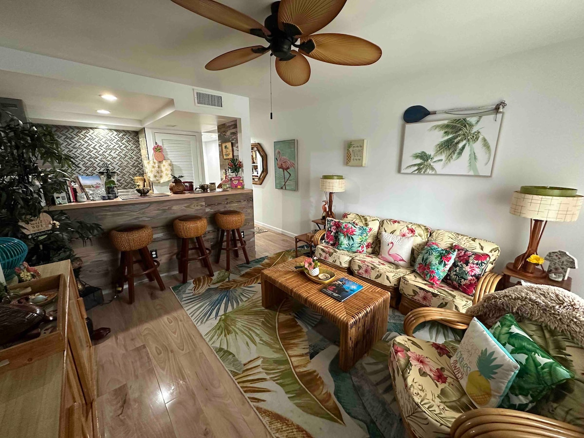 A cozy living room features a bamboo and rattan sofa, adorned with floral cushions. A wooden coffee table holds decorative items. Behind, a kitchen bar with stools provides a casual dining space. Tropical artwork and plants enhance the vibrant ambiance.