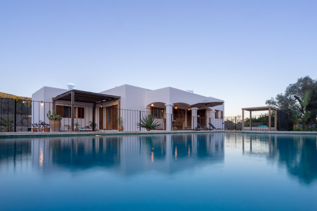 The villa, featuring a modern architectural style, is set against a calm pool that reflects its clean lines. A spacious terrace is visible, accompanied by outdoor seating areas under shaded pergolas, enhancing the serene atmosphere of the surrounding landscape.