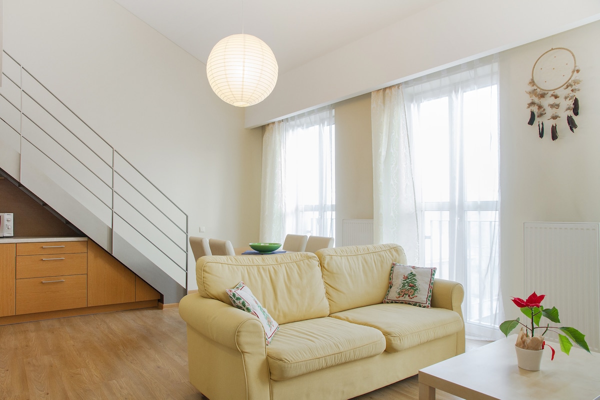 A light-filled living area features a yellow sofa adorned with decorative pillows. Large windows draped with sheer curtains allow natural light to filter in, illuminating the space. A small table holds a potted plant, while a wooden staircase is visible in the background.