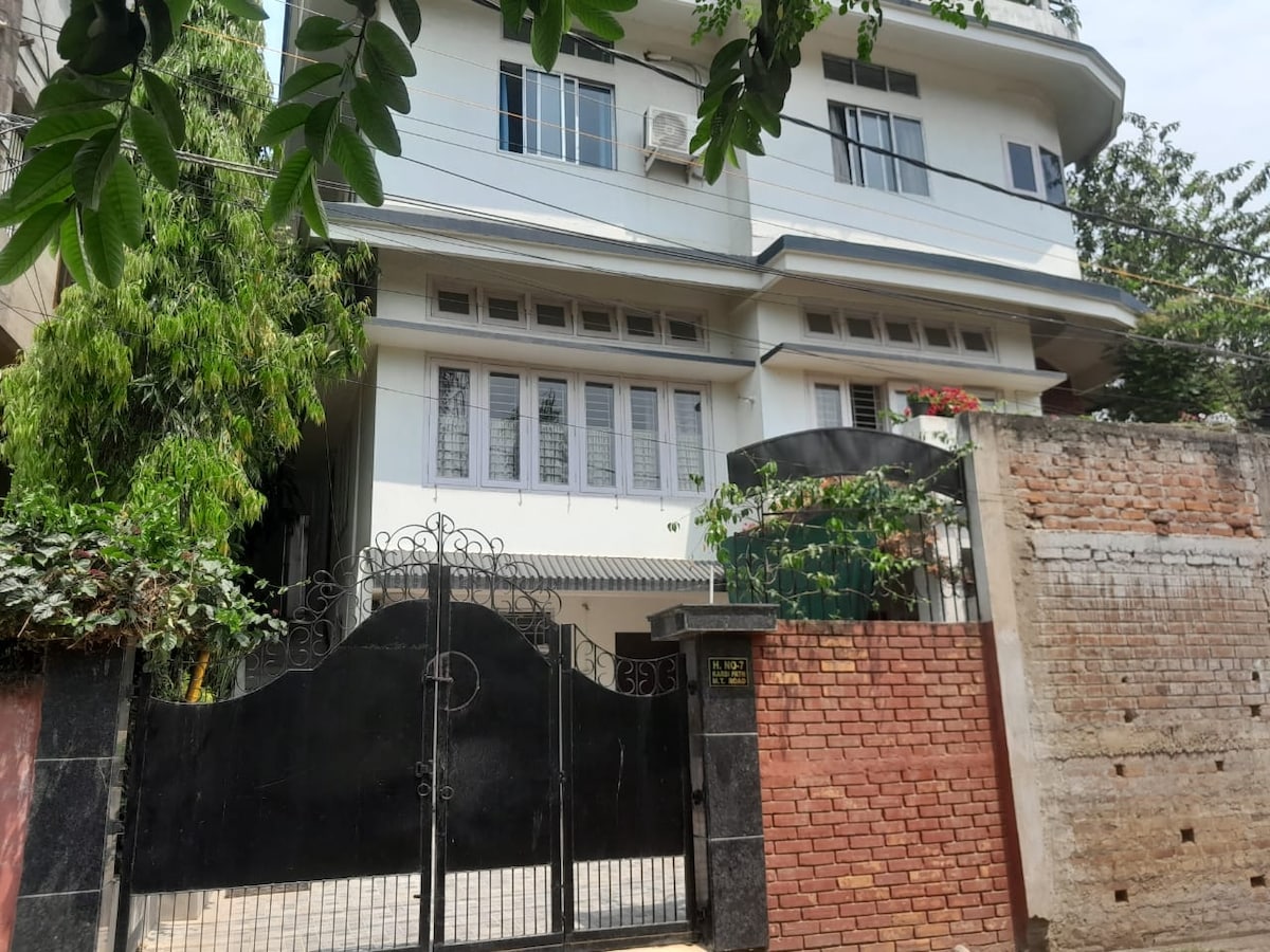 The exterior of the two-story terrace house is presented, featuring a gated entrance with a black ornate gate. The façade is primarily white, complemented by multiple large windows. Lush greenery surrounds the property, enhancing the tranquil environment.