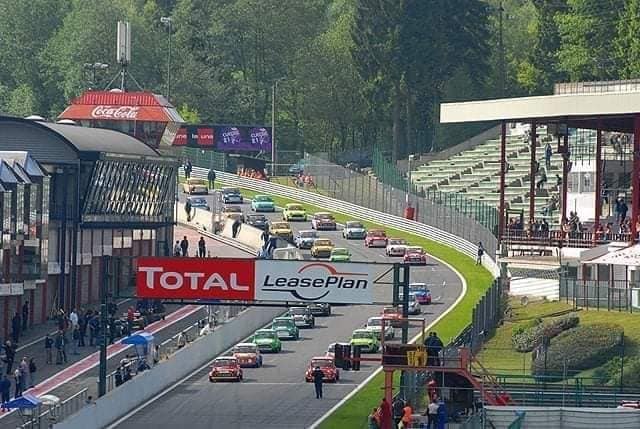 An aerial view captures a race circuit bustling with colorful cars as they navigate the track. Surrounding structures, including grandstands and hospitality areas, are visible, set against a backdrop of lush greenery. Clear signage is prominently displayed, marking the entrance to the circuit.