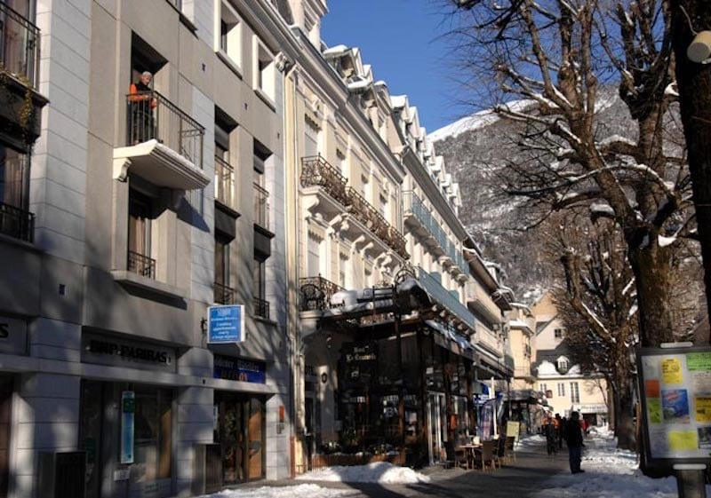 A snow-covered street in a historic city center features elegant buildings with a mix of balconies and windows. Trees line the sidewalk, providing a green contrast against the winter landscape. Pedestrians can be seen walking along the path, adding vitality to the scene.