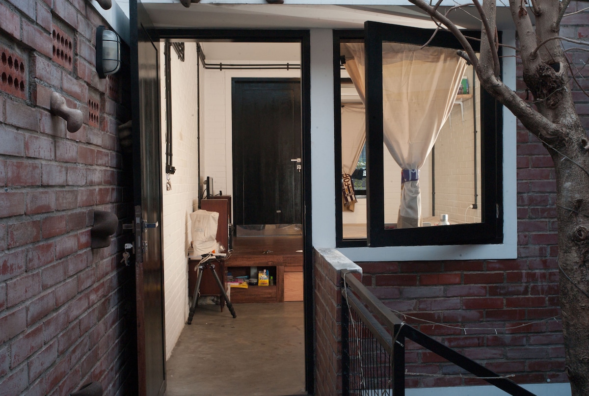 The entrance features a modern design with exposed brick walls and large windows. An inviting view into the interior displays a minimalist decor, with a small seating area and light streaming through the window. A potted tree adds a touch of greenery to the entrance.