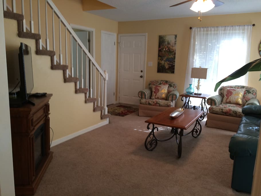 spacious front living area with electric fireplace, tv, ceiling fan and stairs leading to bath and bedrooms.