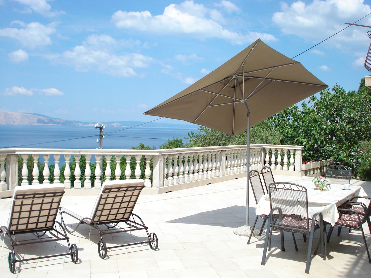 An expansive terrace is showcased with two sun loungers and a large umbrella providing shade. A dining table is set with decorative tableware, offering a view of the sea. Lush greenery borders the terrace, enhancing the scenic surroundings.