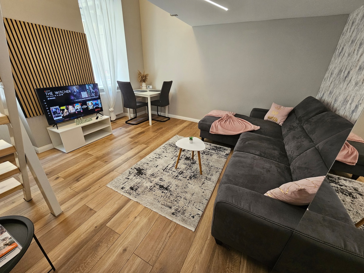 A spacious living area features a large grey sofa adorned with soft pillows, accompanied by a small coffee table. A smart TV is positioned against a textured wall, while a dining table with two chairs is visible in the background. Light streams in through the large window, enhancing the inviting space.