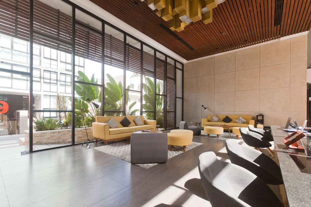 A modern lobby space features large glass windows letting in natural light. Yellow sofas and armchairs are arranged around a circular coffee table. Lush greenery can be seen outside, enhancing the vibrant atmosphere. The ceiling features a unique design, adding character to the space.