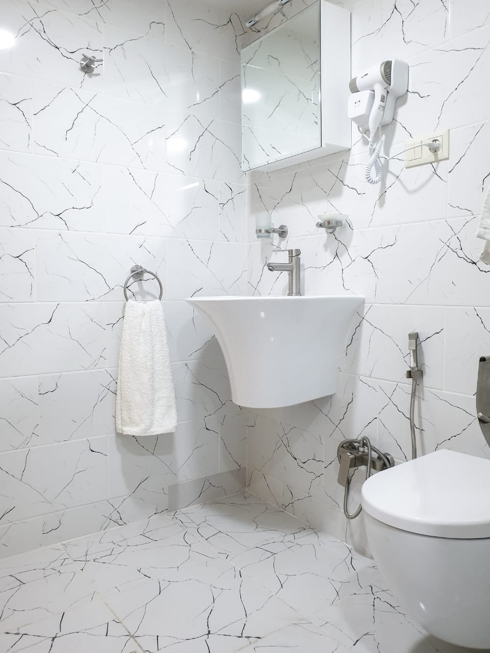 The bathroom features a modern white sink with a sleek faucet mounted on a marble-patterned wall. A large mirror is positioned above the sink, and a towel hangs nearby on a metal holder. A clean toilet is located beside a bidet, enhancing convenience.