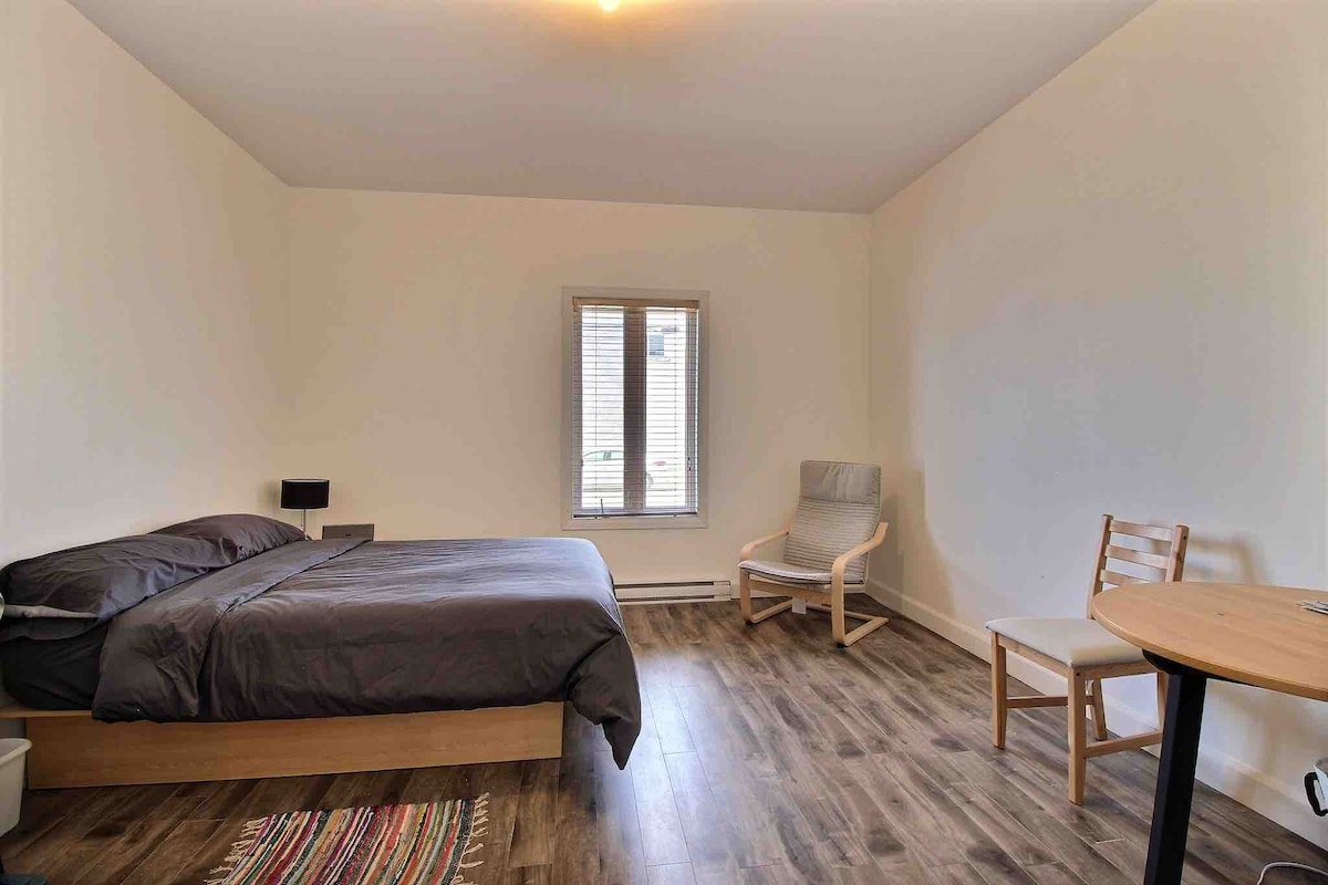 A spacious bedroom features a comfortable double bed dressed in dark bedding. A small window allows natural light to enter, and a cozy chair is positioned nearby. A wooden table and chair are visible, providing a simple workspace. Wooden flooring enhances the room's warmth.