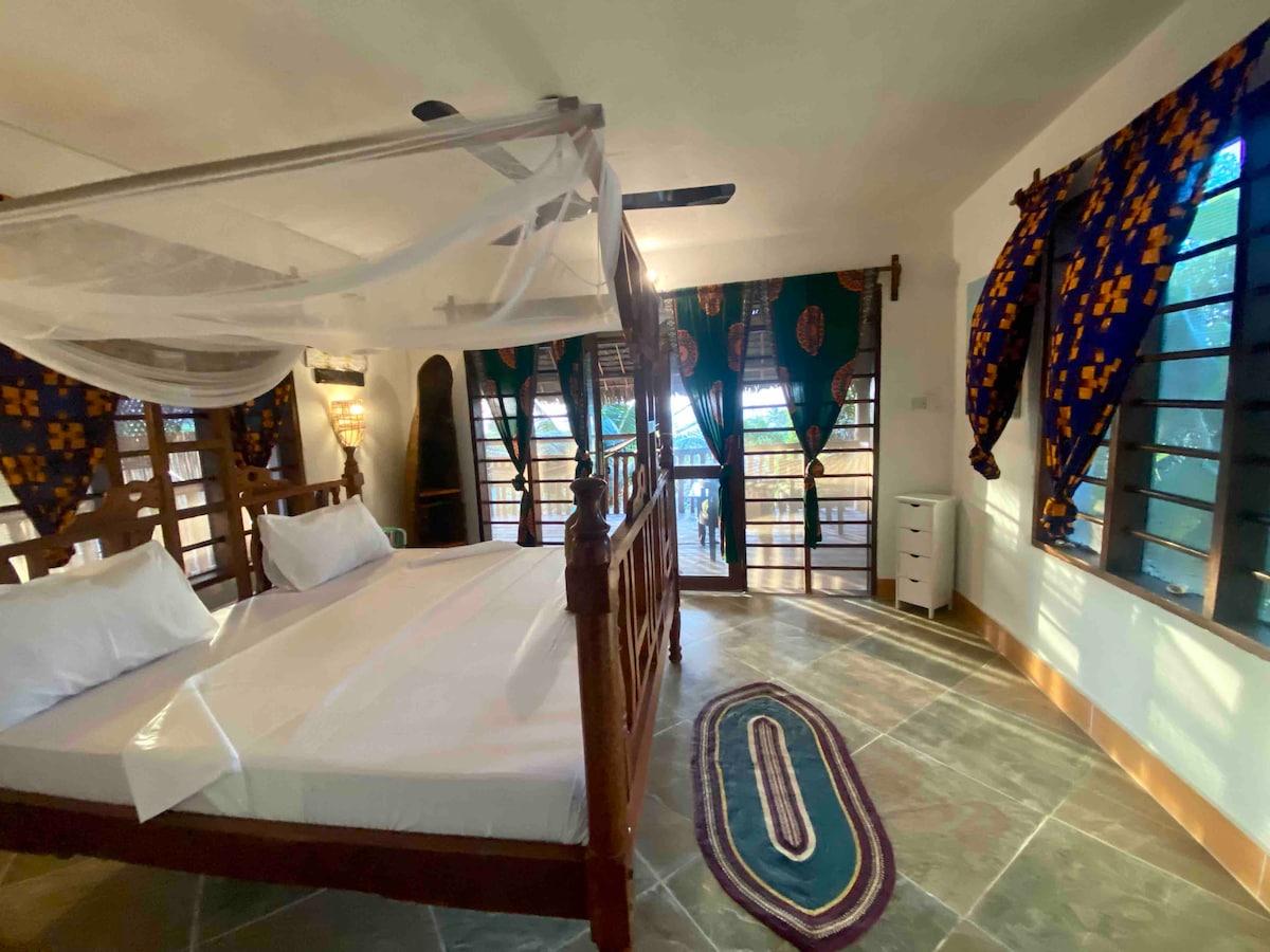 A spacious room features a wooden four-poster bed with a mosquito net. Colorful patterned curtains frame the windows, allowing natural light to illuminate the stone floor. A small white cupboard is positioned near the entrance, enhancing the room's functionality.