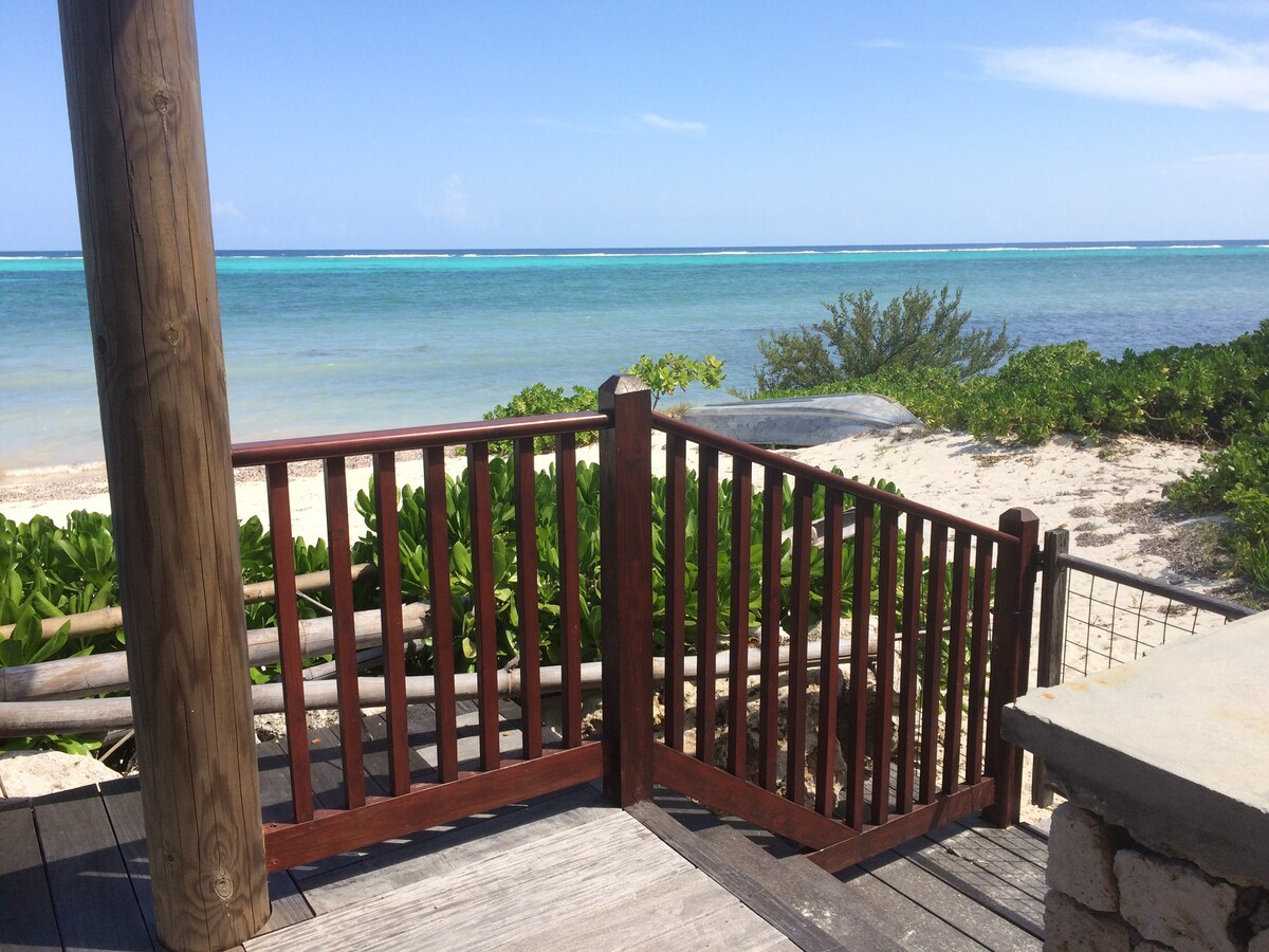 A wooden gate leads to a sandy beach area, with the clear turquoise waters of the ocean visible in the background. Lush green vegetation edges the beach, creating a natural boundary. The sky above is bright and cloudless, enhancing the sense of tranquility.