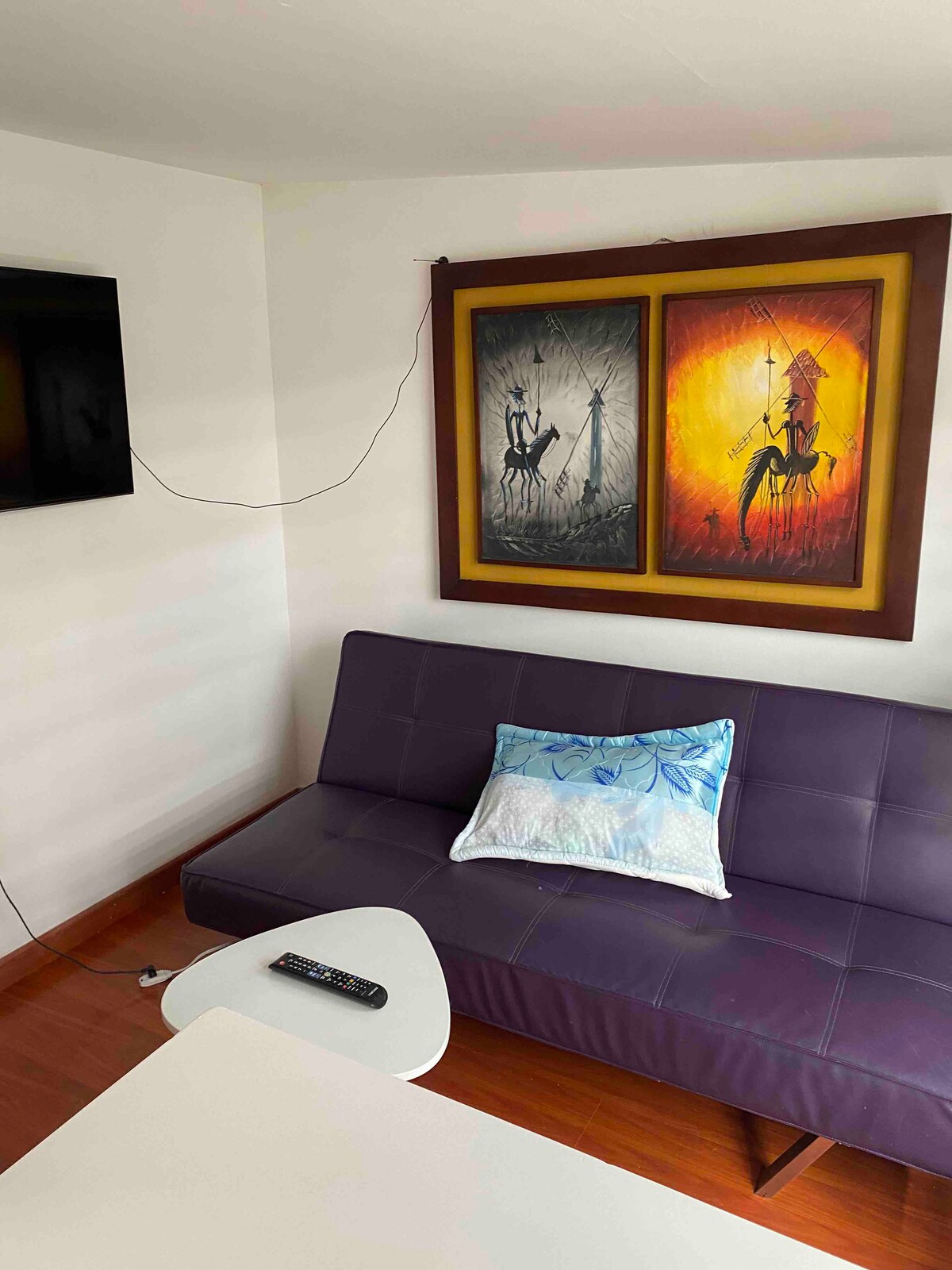A modern seating area features a purple sofa with a decorative pillow, positioned next to a sleek white table. Two vibrant framed artworks adorn the wall, creating a focal point, while a television is mounted opposite the seating area.