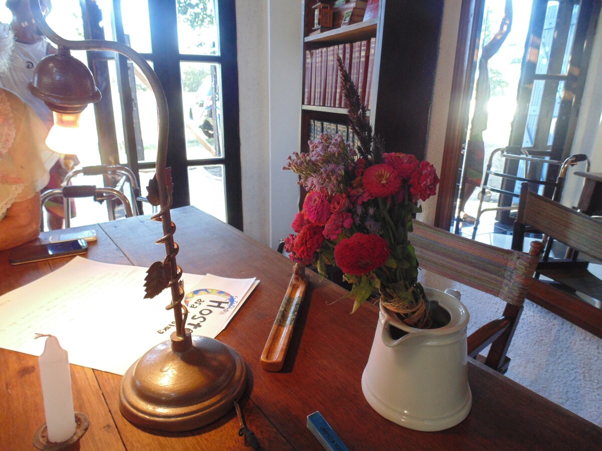 A rustic wooden table is adorned with a bouquet of colorful flowers in a ceramic vase. A brass desk lamp provides soft lighting, while sunlight filters through multiple glass doors in the background, enhancing the inviting ambiance of the space.
