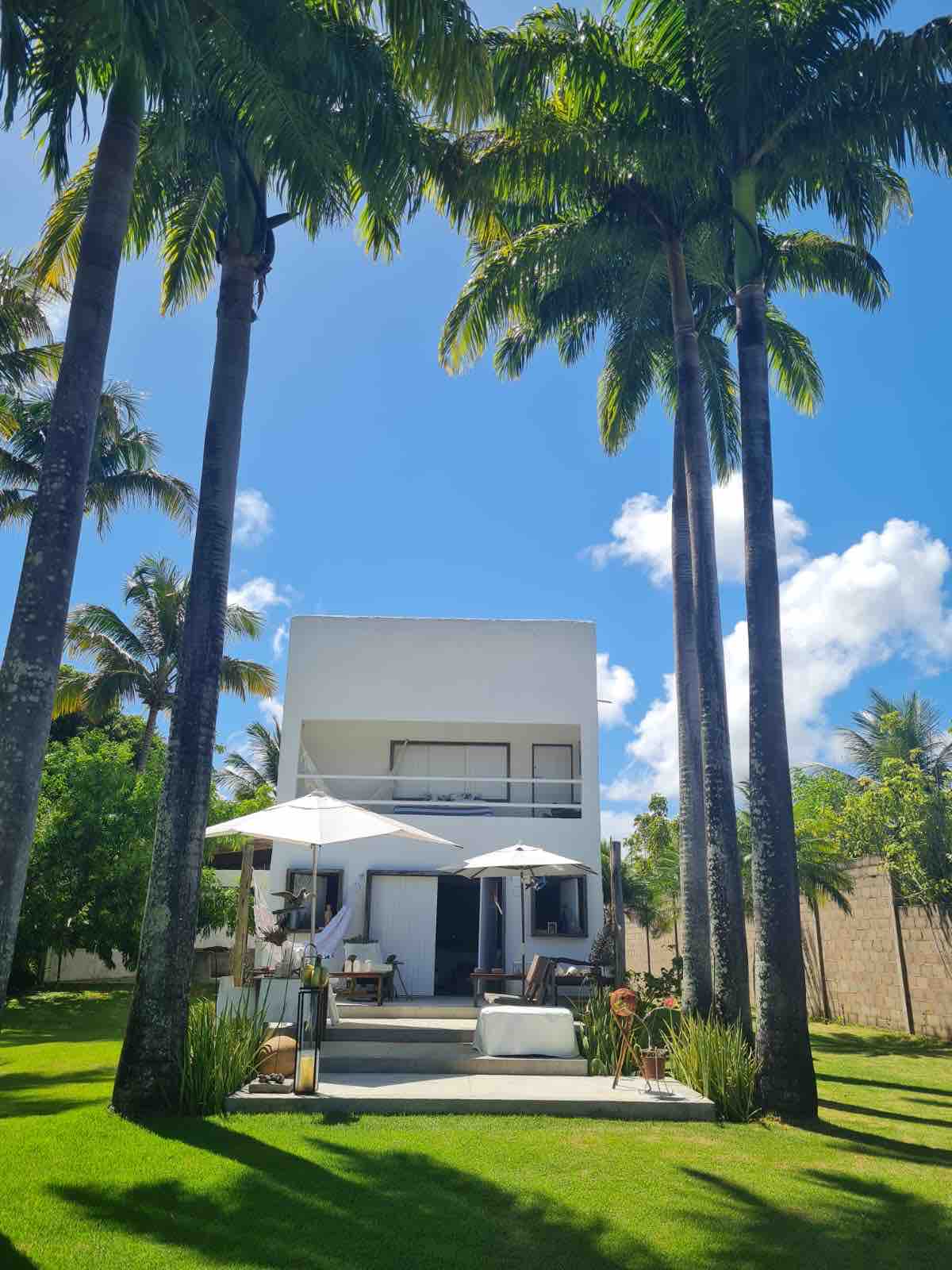 A modern two-story house is framed by tall palm trees under a clear blue sky. The exterior features a light-colored facade and spacious patio area with lounge chairs and umbrellas, providing a relaxed outdoor setting.