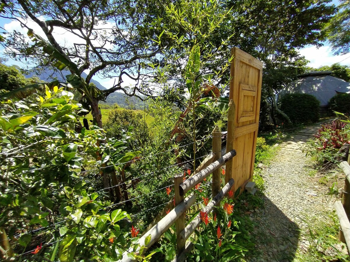 A wooden door opens onto a meandering path surrounded by lush greenery and colorful flowers. The flourishing landscape includes trees and plants, enhancing the natural setting. Mountains are faintly visible in the background, contributing to the serene atmosphere of the area.