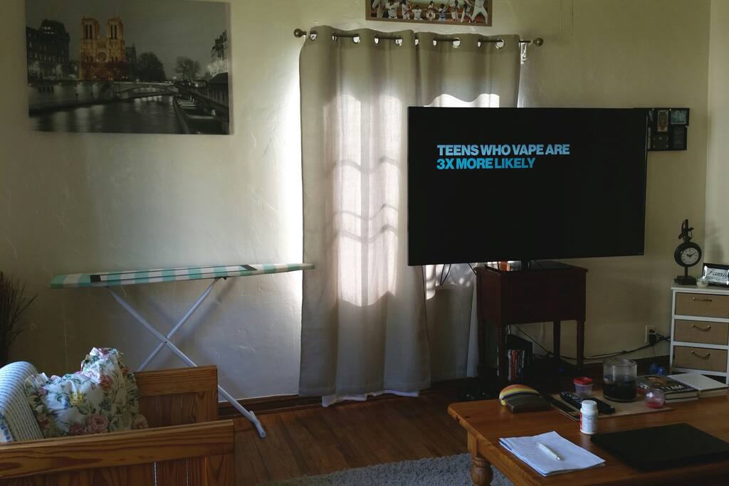 Different angle of the living room. Contains 1 full couch, single chair, white shag rug, coffee table, iron/ironing board, 72" LED Smart TV with cable and wifi.