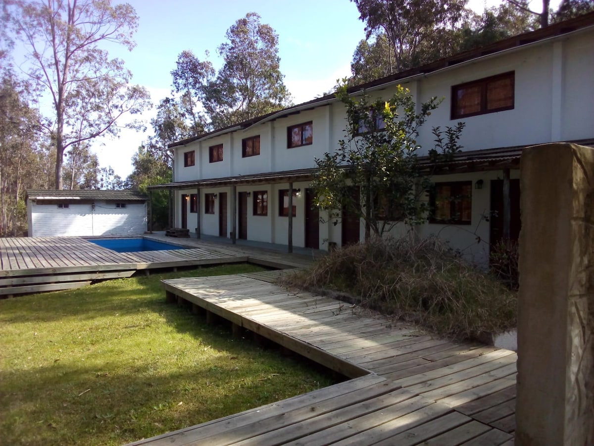A two-story building is surrounded by trees and natural foliage. A wooden deck leads to the entrance, while a pool is partially visible in the background. The exterior features multiple windows and doors, allowing for ample natural light.