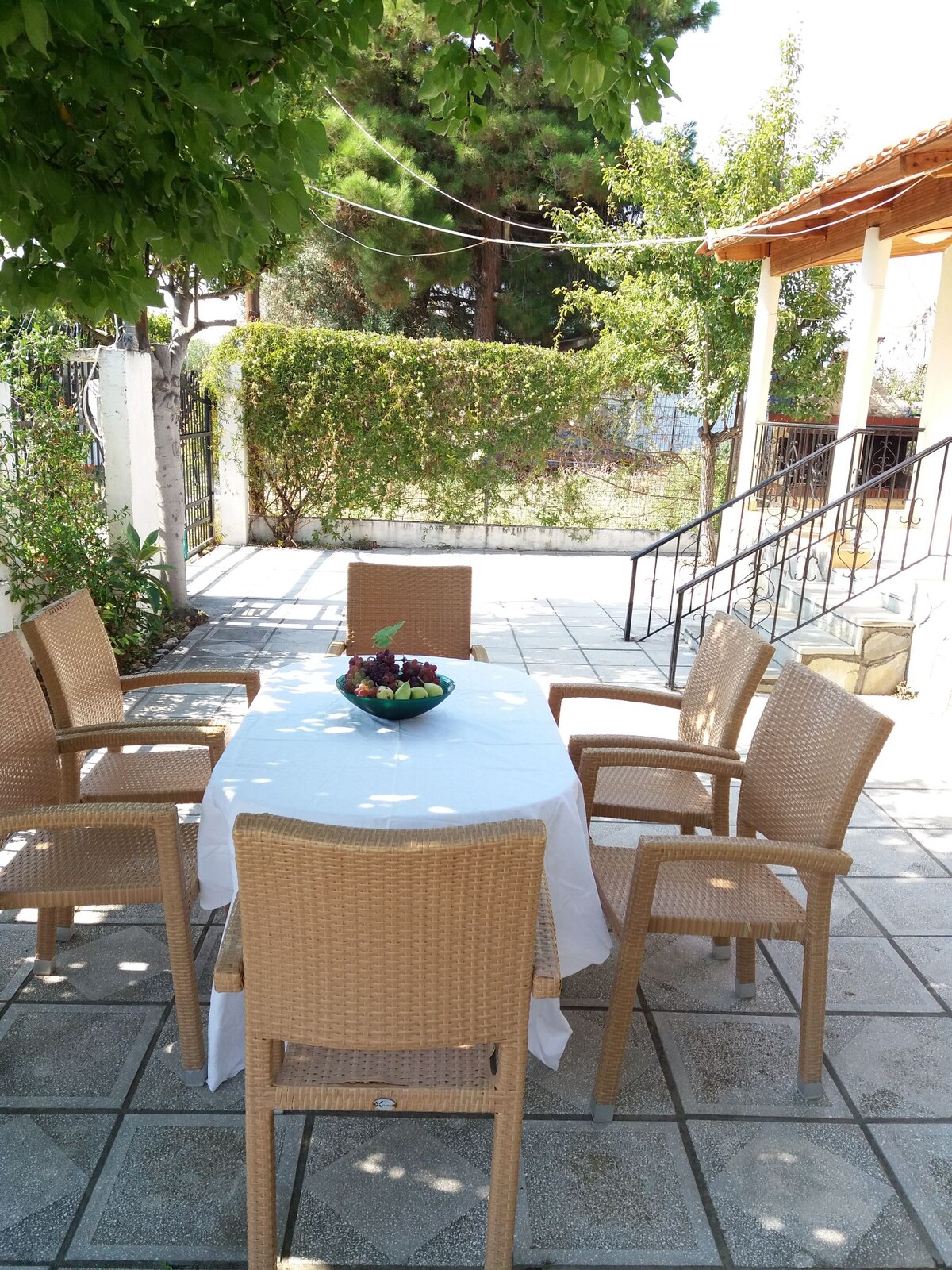 An outdoor dining area features a round table set with a fruit bowl, surrounded by six wicker chairs. The space is shaded by a leafy tree, with a stone pathway leading to a set of stairs that provides access to the house.
