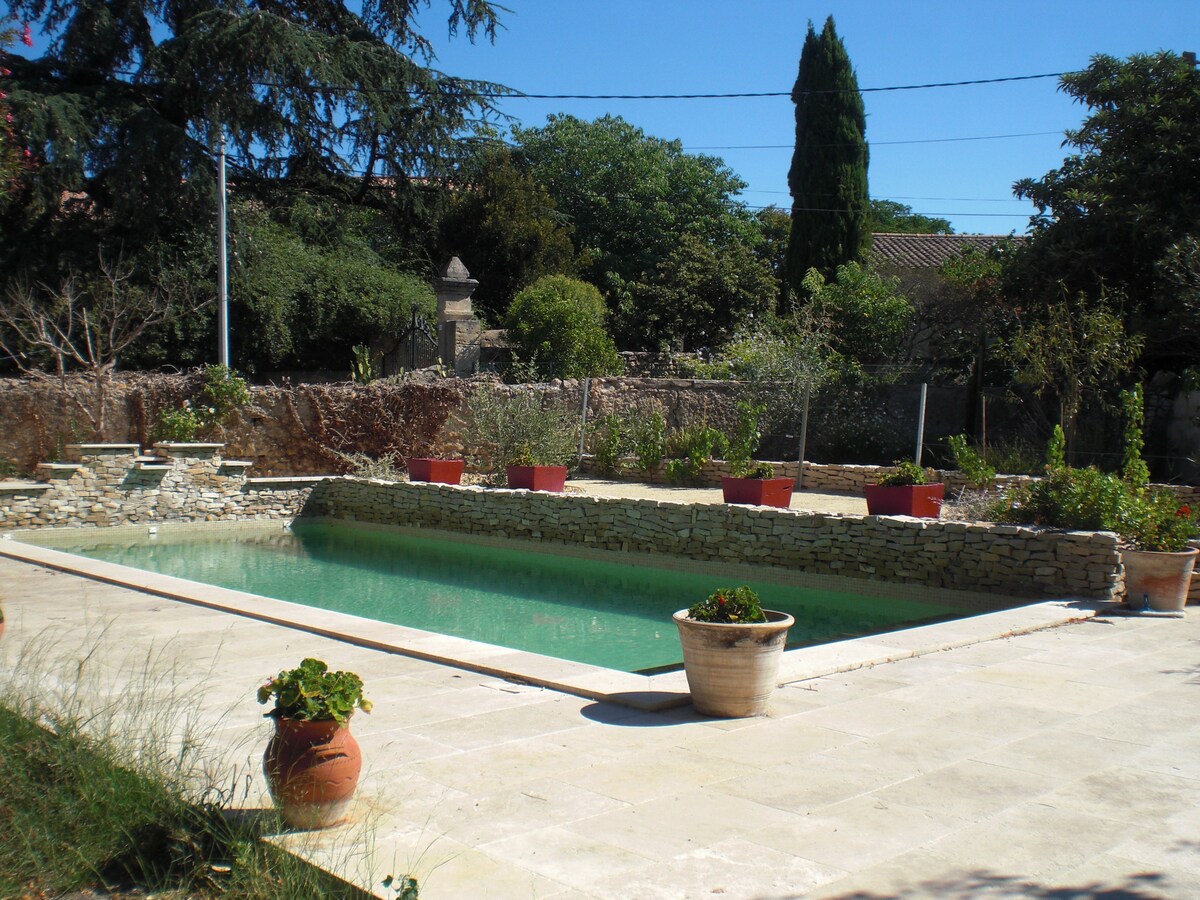 A private swimming pool is surrounded by a stone patio, featuring lush greenery and potted plants placed around the perimeter. The tranquil water reflects the clear blue sky, while trees and shrubs create a natural backdrop. The setting invites relaxation and enjoyment in the sun.