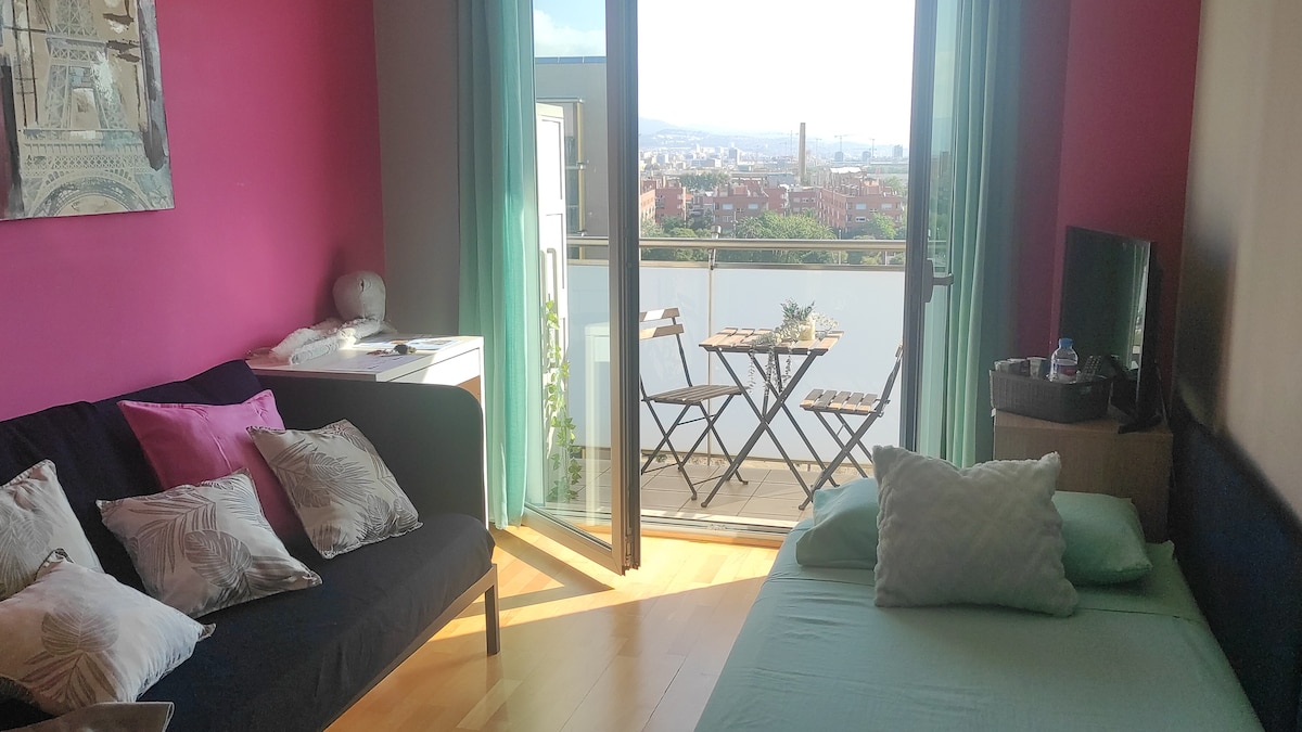 The bright room features a black sofa and a bed with light-colored linens. Large glass doors open to a small balcony with seating. Pink walls and wooden flooring enhance the space, with soft natural light illuminating the area from the outdoor views.