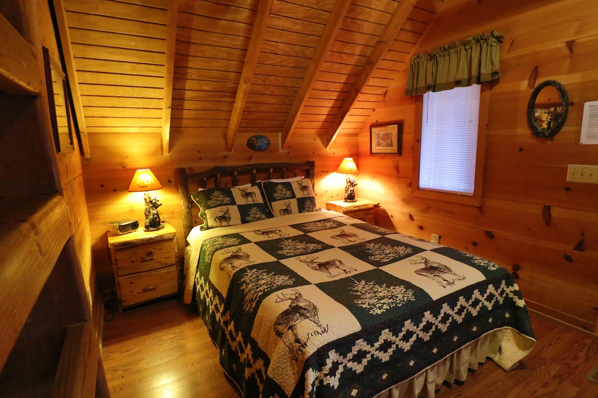 A cozy bedroom features wood-paneled walls and a vaulted ceiling with exposed beams. A spacious bed is adorned with a quilt displaying rustic patterns. Two bedside tables with lamps are positioned on either side. Natural light enters through a window dressed with green curtains.