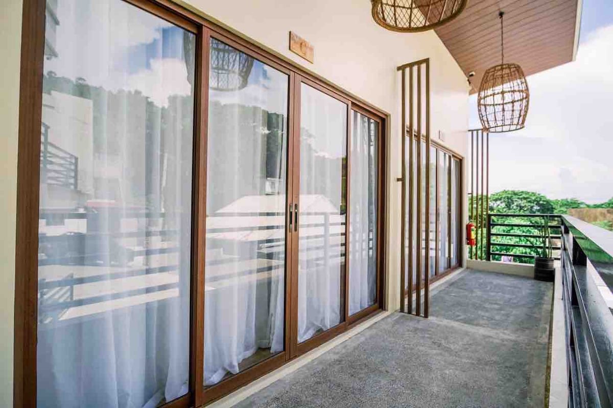 A spacious balcony features sliding glass doors framed in warm wood, allowing natural light to fill the area. Soft white curtains gently flutter in the breeze. Two hanging light fixtures add a touch of style, and a railing offers an unobstructed view of the surrounding greenery.