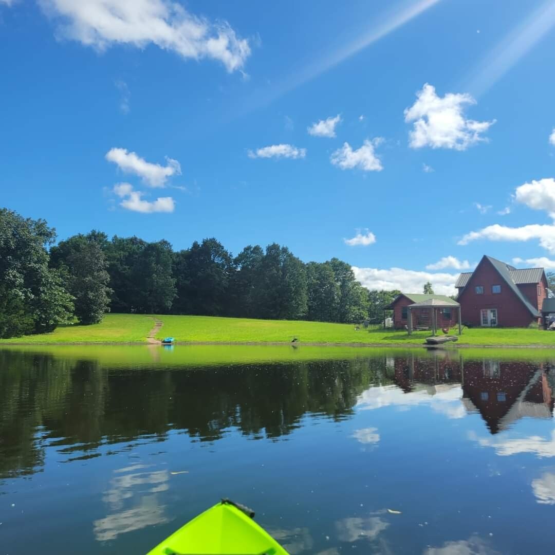 A serene landscape is depicted, featuring a calm pond reflecting blue skies and surrounding greenery. A well-maintained lawn leads to a charming red house in the background, complemented by a few trees. The image captures a peaceful atmosphere ideal for outdoor activities.