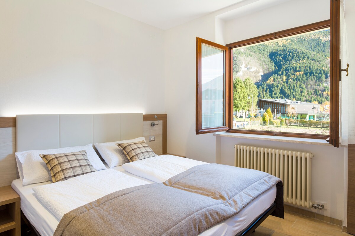 A well-lit sleeping area features a spacious bed with neutral bedding and decorative plaid throw pillows. A large window offers a view of the surrounding mountains and greenery. Natural light enhances the calming ambiance of the room.