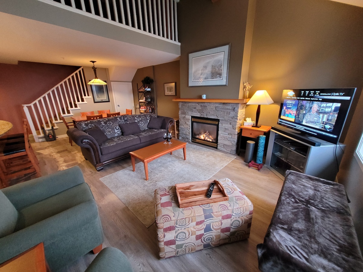 A spacious living area features a stone fireplace framed by a wooden mantle, with a comfortable brown couch and armchair arrangement. A flat-screen TV is positioned nearby, and a large coffee table sits on a textured rug. The dining area is visible in the background.