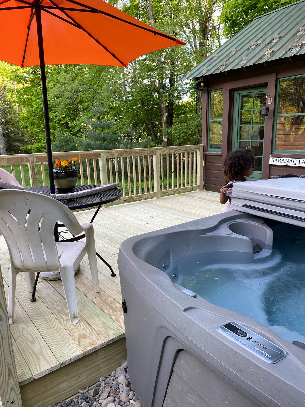 An outdoor deck features a hot tub adjacent to a small table with chairs. A bright orange umbrella provides shade. The lush greenery of the surrounding forest is visible in the background, enhancing the tranquil outdoor setting.