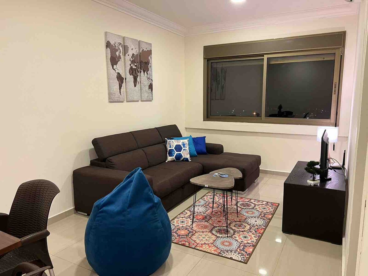 A living room features a spacious sectional sofa adorned with colorful cushions. A small coffee table is positioned on an area rug, adding warmth. A large window allows for natural light, while wall art depicting a world map enhances the decor.