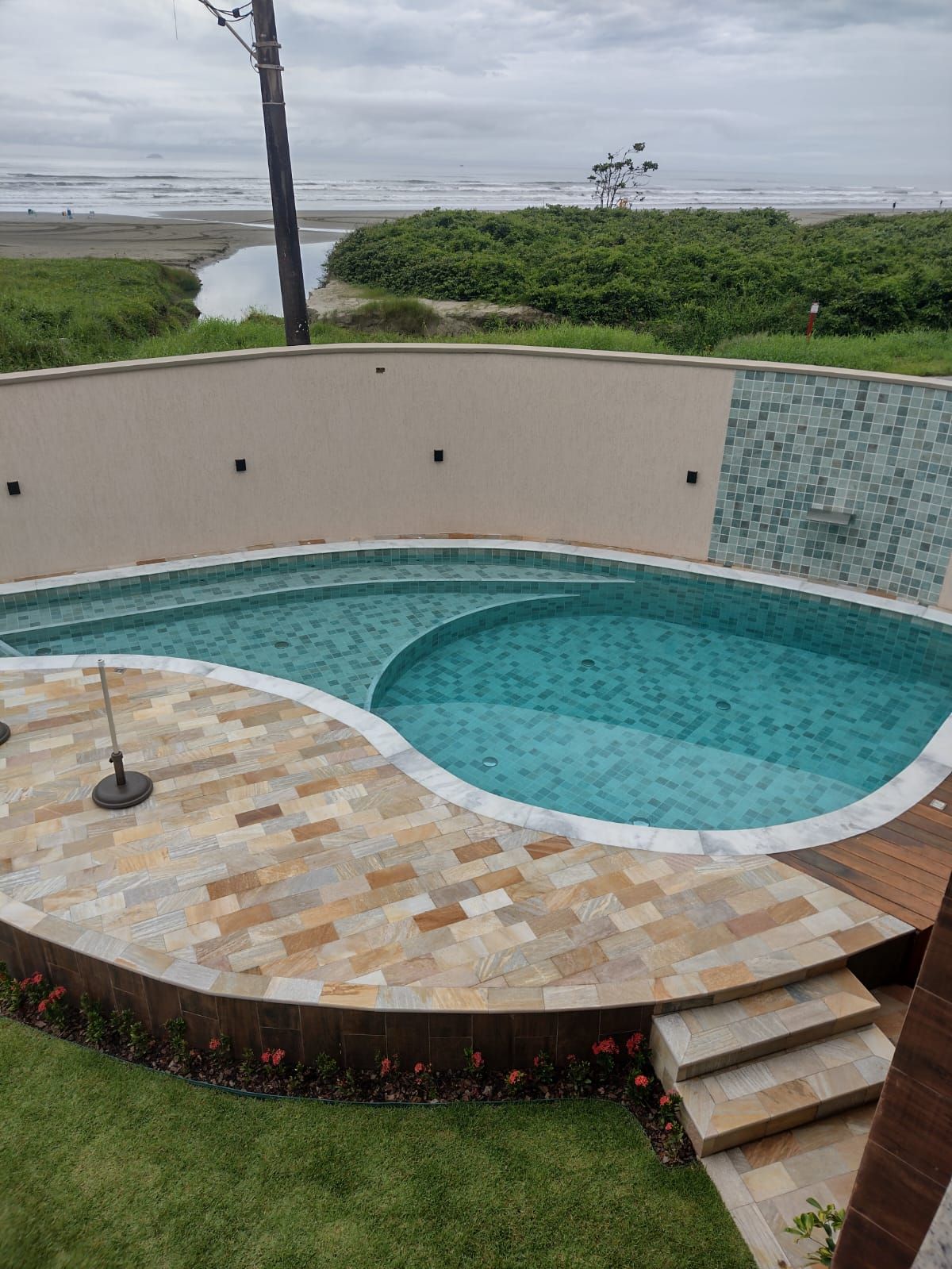 A modern heated swimming pool features a unique design with smooth curves, surrounded by light-colored stone tiles. Lush greenery outlines the area, and a shallow section is visible. The pool area is complemented by a wall with mosaic tiles, creating an inviting space.
