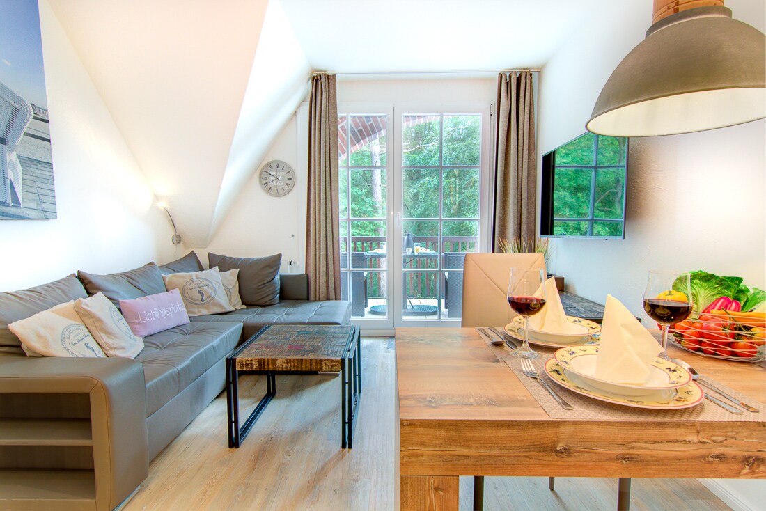 A modern living area is presented with a comfortable sofa and a coffee table. A dining table is set with plates and wine glasses. Natural light enters through large windows, leading to a balcony with outdoor views. Neutral tones create a welcoming ambiance.