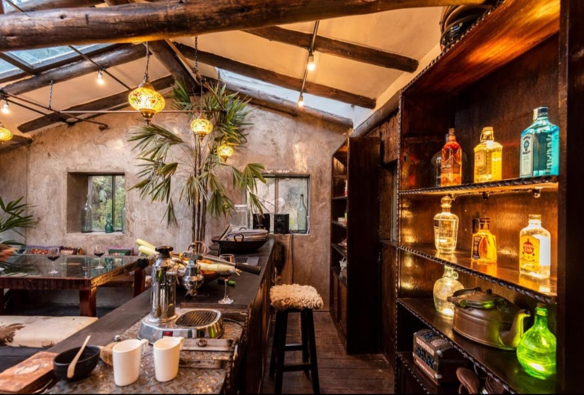 A rustic kitchen area features a wooden bar with a variety of colorful bottles neatly arranged on shelves. Ambient lighting from pendant lamps highlights the natural textures of the walls and ceiling beams. Fresh greenery can be seen through the window, adding a lively touch.