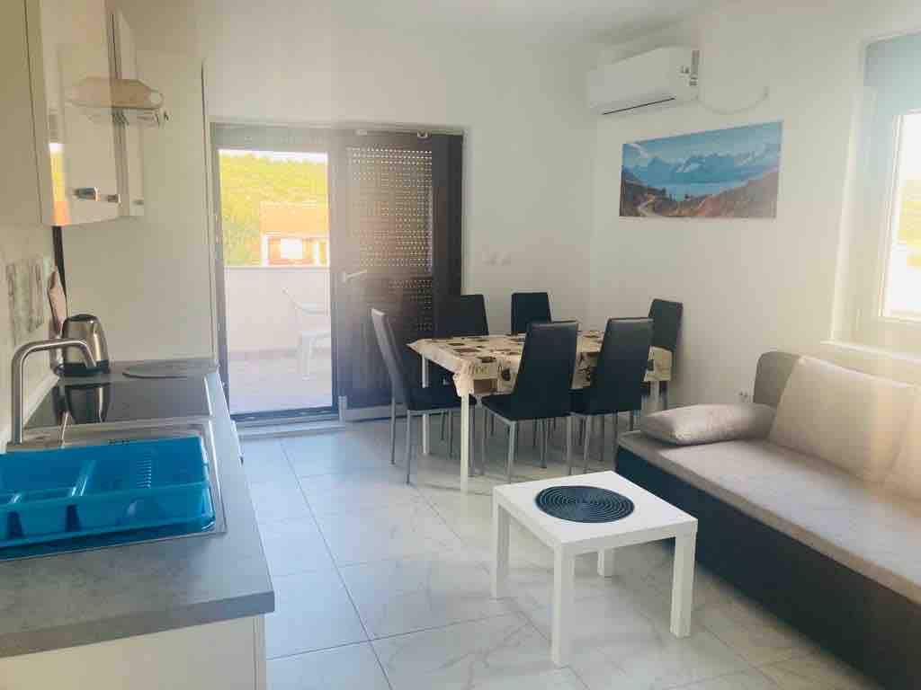 A bright living and dining area is visible, featuring a table with six chairs and a relaxing sofa. Large windows provide natural light, and a door leads to the terrace. The kitchen area includes modern cabinetry and a sink, with a blue dish rack placed on the countertop.