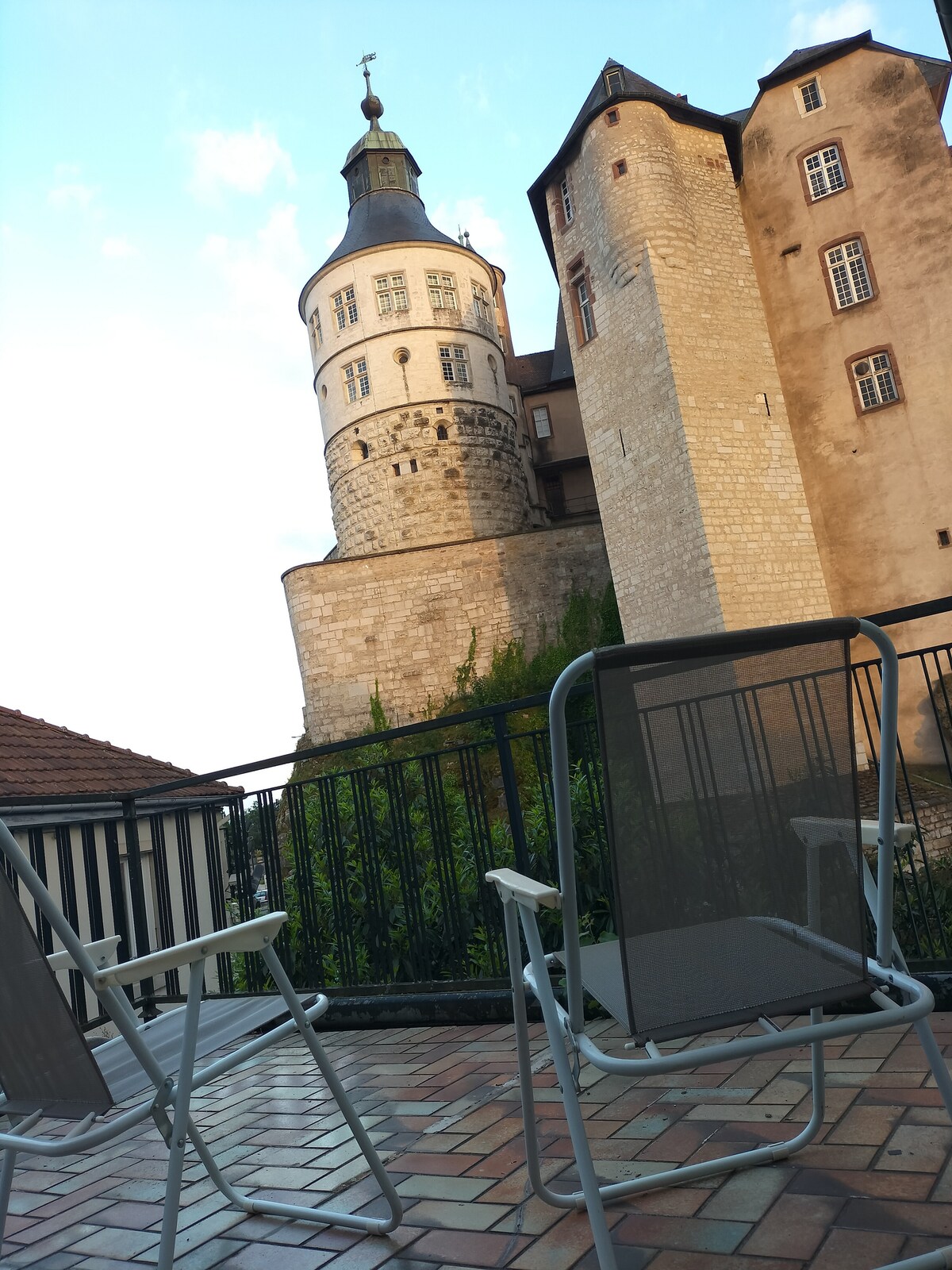 A view from a terrace displays a castle with a mixture of stone and brick walls, including a prominent turret. Two lounge chairs are positioned on a tiled surface, offering a spot for relaxation while enjoying the scenic landscape.