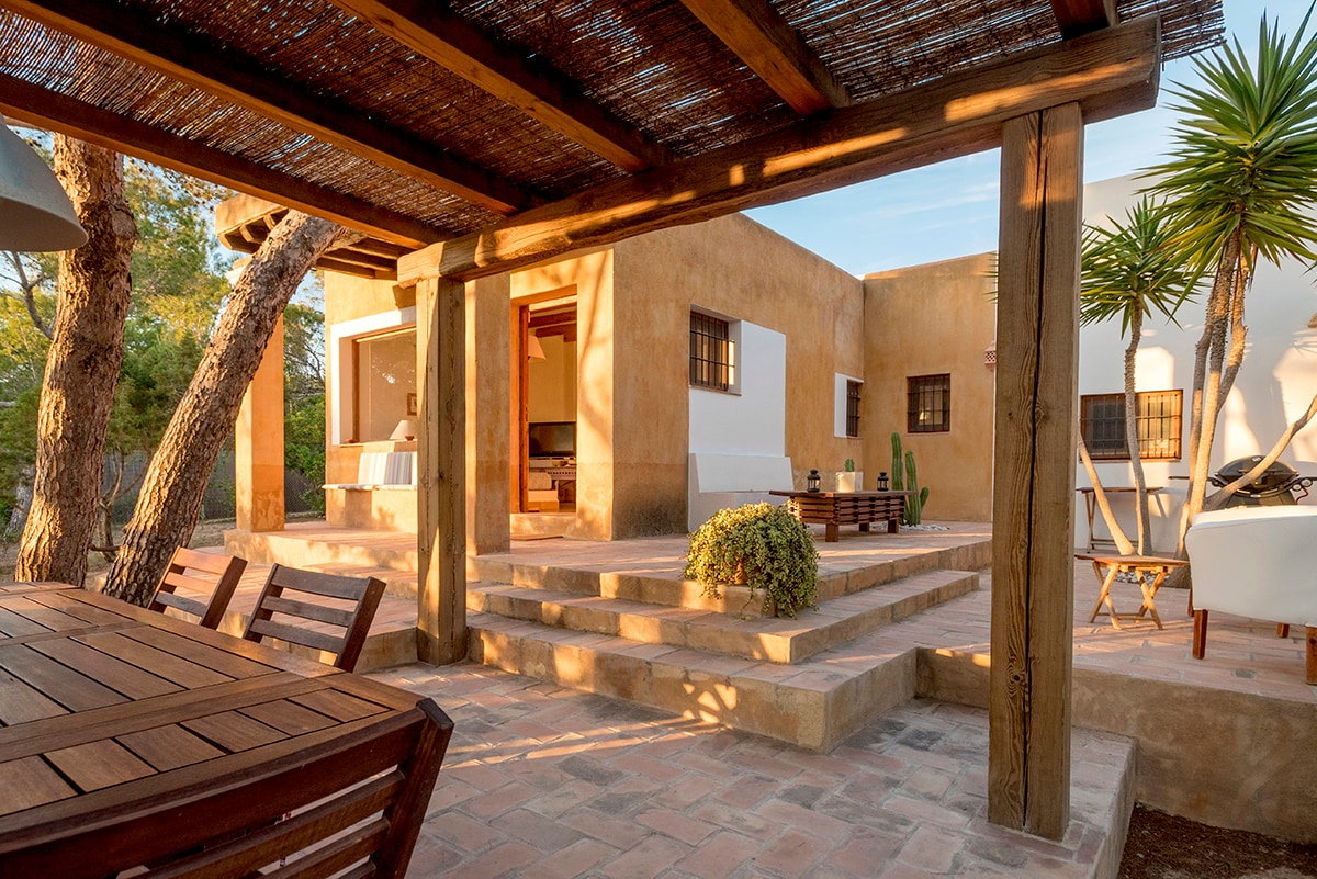 An outdoor seating area features a wooden table and chairs shaded by a pergola. The setting includes natural stone steps leading to a rustic-style house with large windows. Lush greenery and decorative plants enhance the tranquil atmosphere.